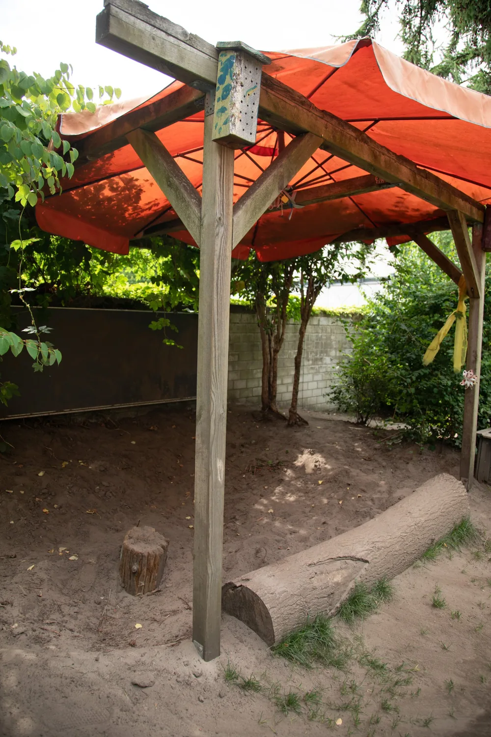 Pavillon mit orangem Sonnensegel, Baumstamm und Sandfläche im Garten der Kita